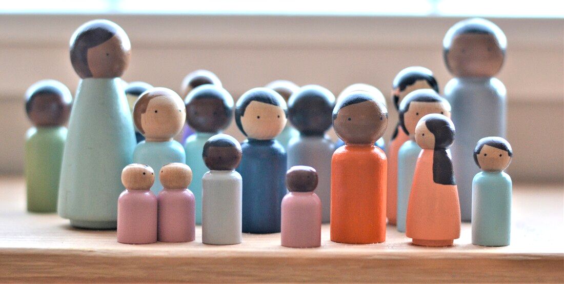 A group of small wooden peg dolls in soft colors and varied skin tones, gathered together on a sunlit wooden shelf